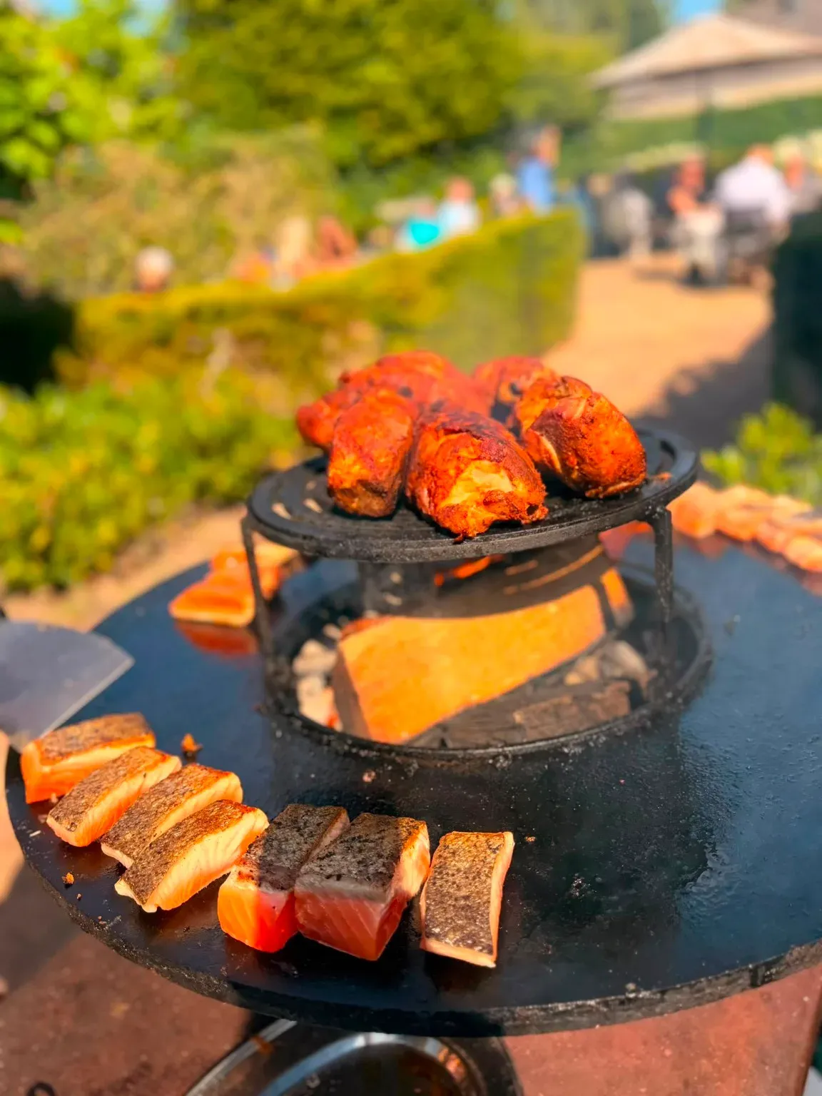 Ofyr met gemarineerde kip en zalm van de plancha, in de tuin van Het Oude Raadhuis met gasten op de achtergrond