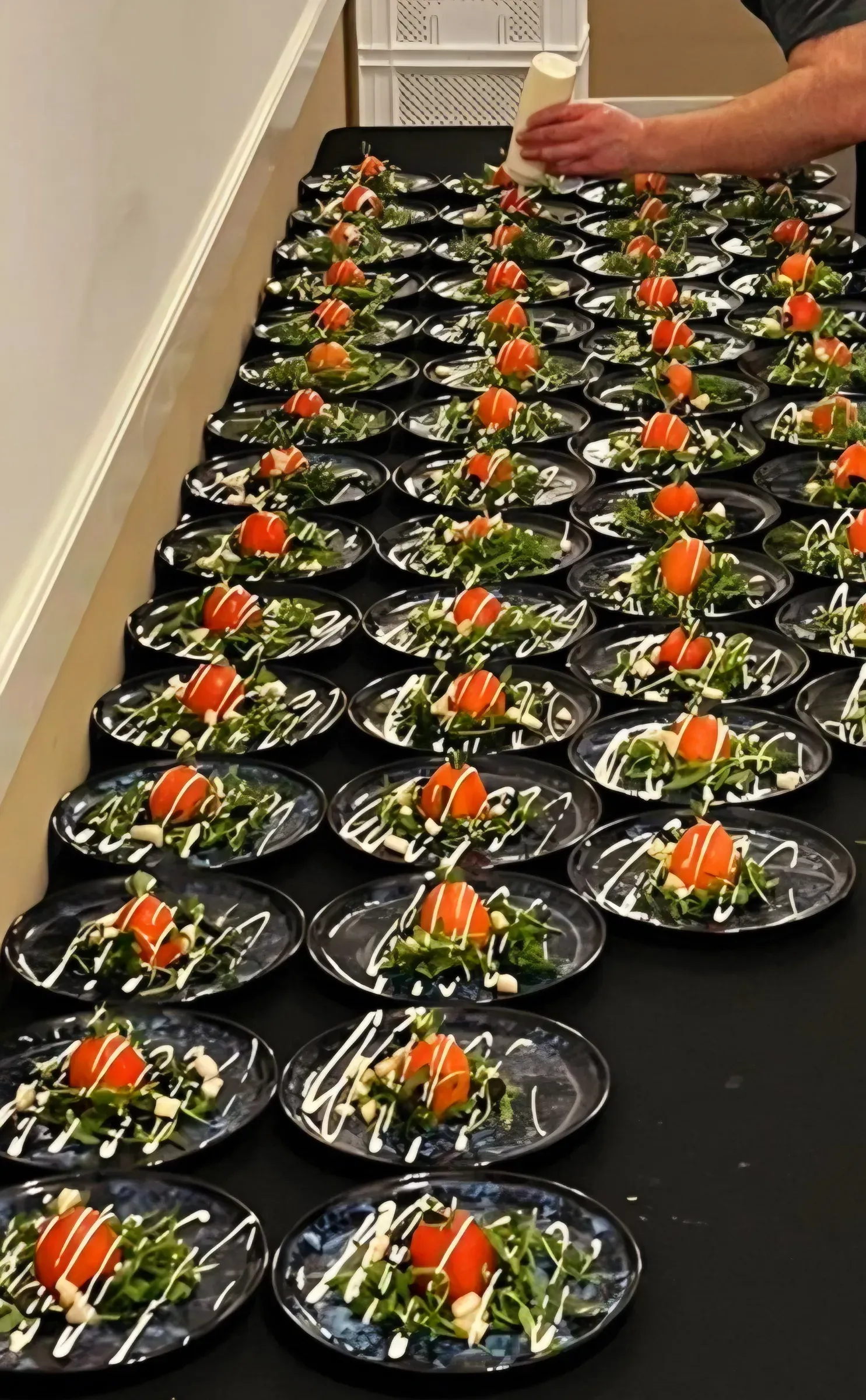 Lange rij zwarte bordjes met een hapje van groen, crème en tomaat, klaar voor uitgifte